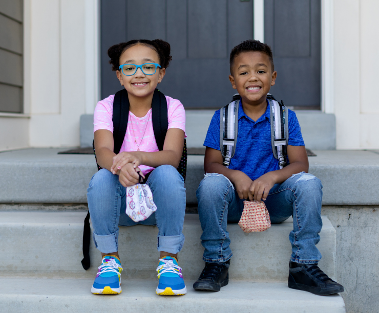 Siblings sit outside their home holding homemade masks