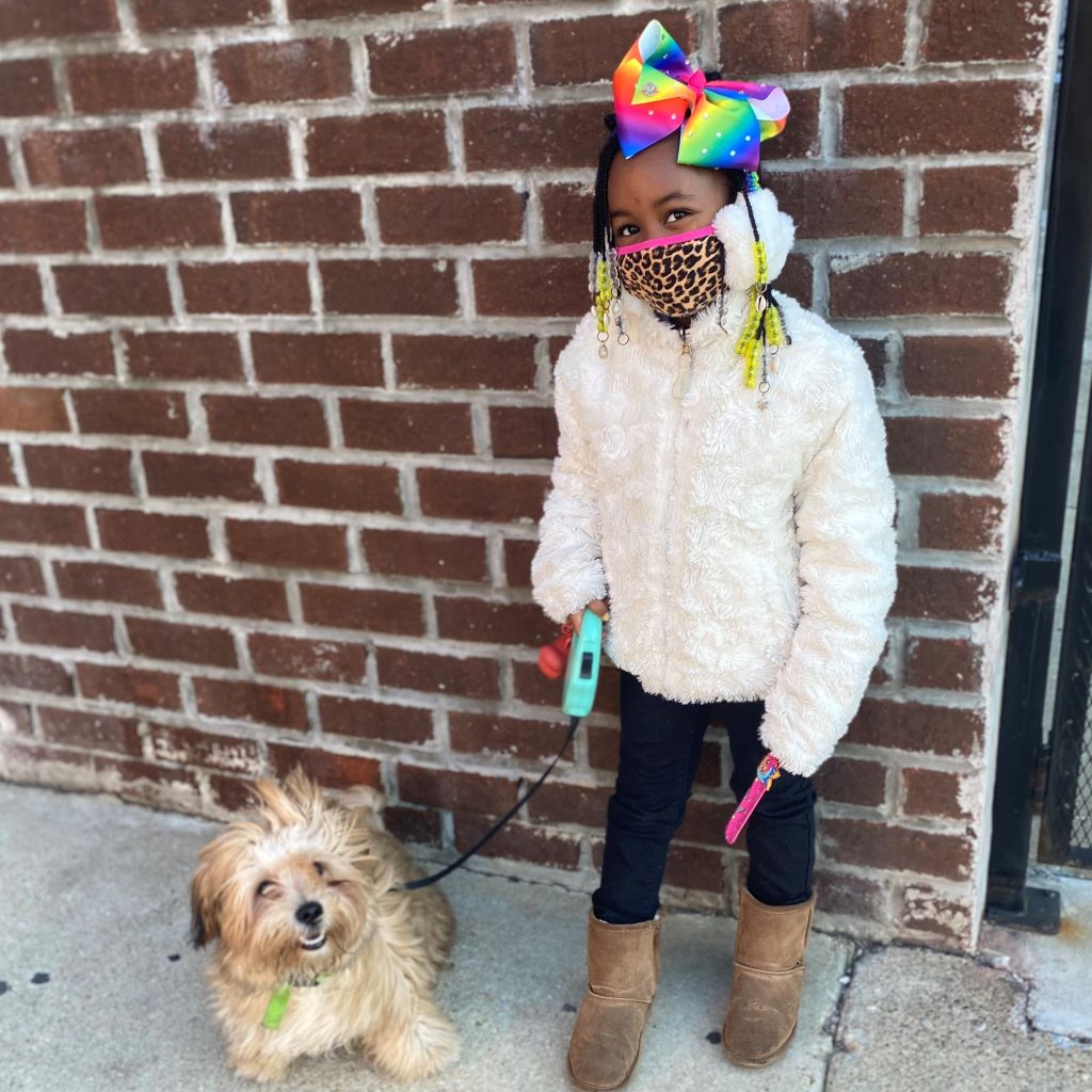 Child with rainbow hair bow and cheetah print mask poses while holding a small dog on a leash.