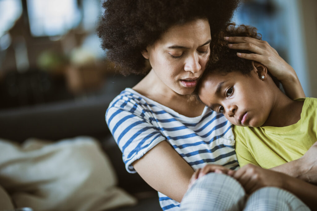 Mother comforting elementary-age daughter, hugging and sitting side-by-side in front of a couch.