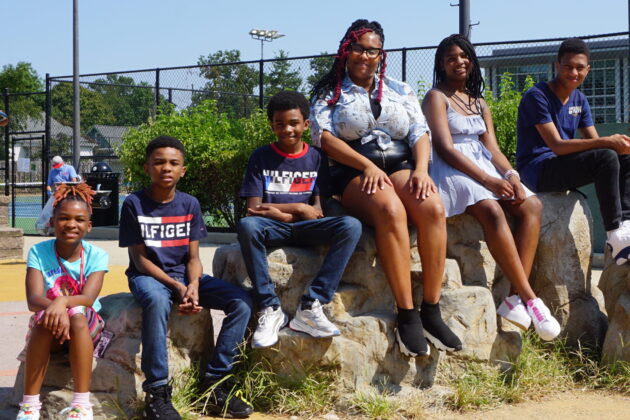 Mother with five pre-teen and teen children sitting on rock formation at playground smiling for the camera.