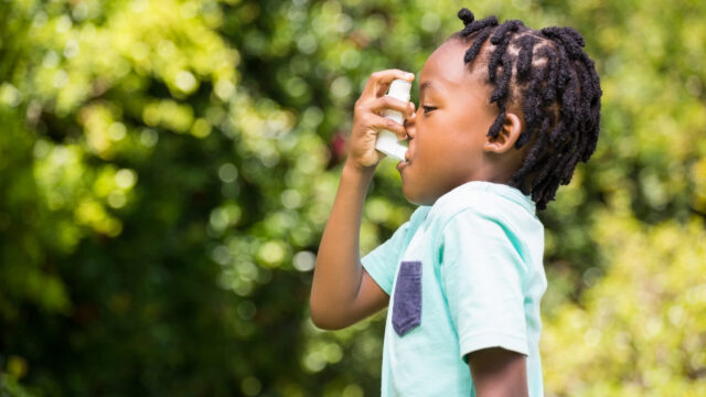 Photo of side profile of boy using an asthma inhaler in a park.