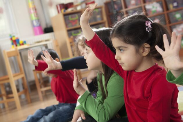 Several young children in a classroom sitting on the floor and raising their hands.