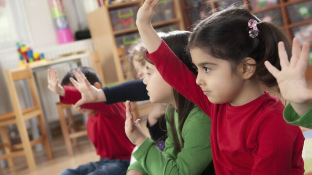 Several young children in a classroom sitting on the floor and raising their hands