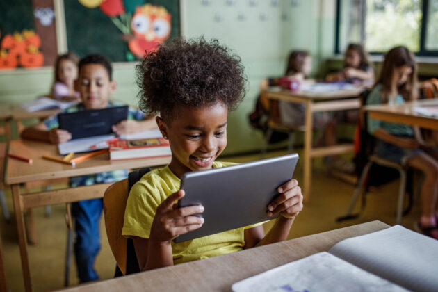 Black elementary student smiling in classroom, sitting at a desk and looking at a handheld electronic tablet.