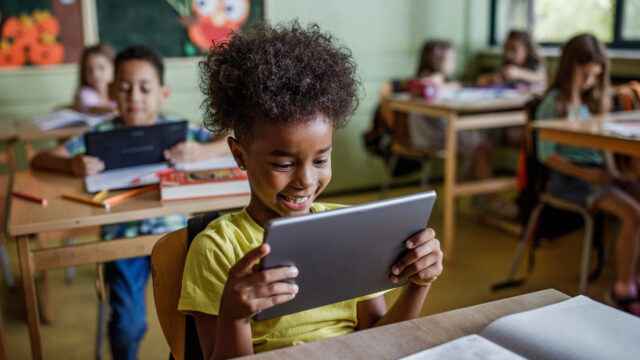 Black elementary student smiling in classroom, sitting at a desk and looking at a handheld electronic tablet.