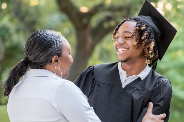 Photo of woman in white shirt embracing smiling young man in black graduation robe and mortarboard. They are outside, and a tree is visible in the background above them.