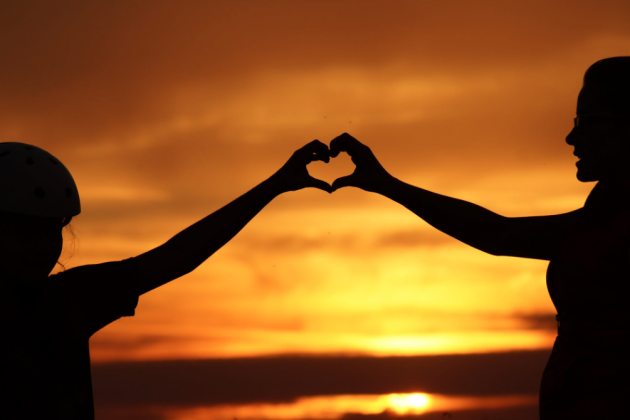 Stock photo of people making a heart shape with their hands in front of a sunset.