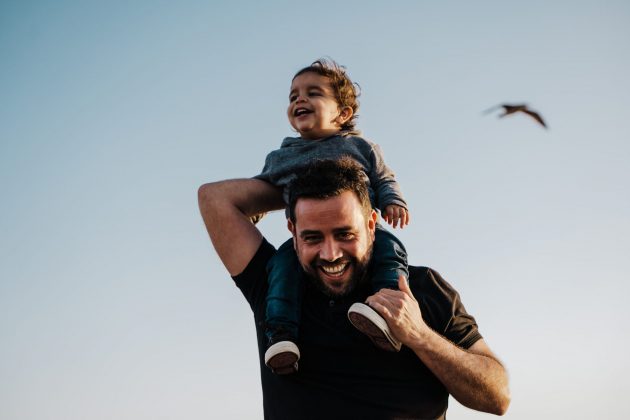 Young child on man's shoulders, both smiling. Bird flies behind them in the distance.